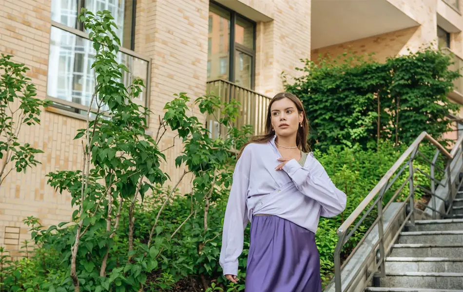 Kvinne står ute i en trapp foran grønne vekster iført lilla skjørt og dus lilla bluse. 