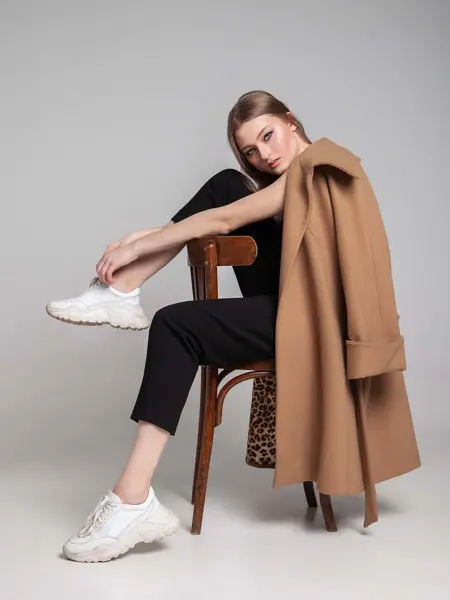 Kvinne med hvite, chunky sneakers som sitter bak-fram på en stol med beige jakke hengende over skuldrene. Illustrasjonsbilde til artikkel om vårmote for sko.