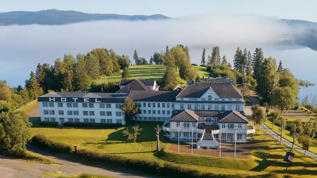 Sommerlig bilde av fasaden på hotellet med sol, grønt gress og tåke i bakgrunnen