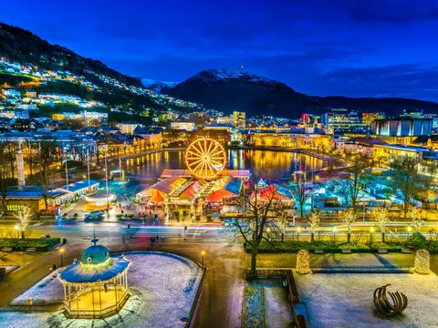 Julemarkedet i Bergen ved Lille Lungegårdsvann sett ovenfra en kveld, med opplyst pariserhjul, musikkpaviljongen og snødekte Ulriken bak.