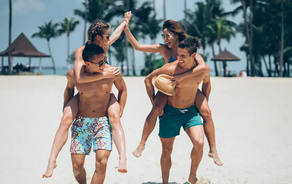 To menn med mønstrete og grønn badebukse bærer to damer på ryggen på en strand  