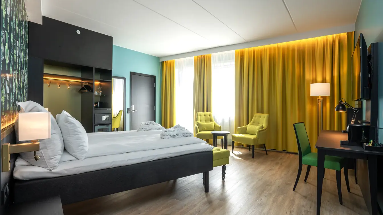 Gule gardiner, to gule lenestoler, salongbord, skrivebord, grønn stol, TV, vindu, gule gardiner, veggspeil, dobbeltseng.