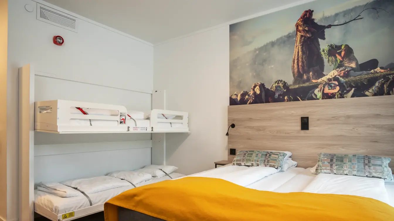 Familierom med oppredde køyesenger til venstre og dobbeltseng til høyre. Over sengegavl henger et bilde fra en Peer Gynt-forestilling. Parkettgulv.