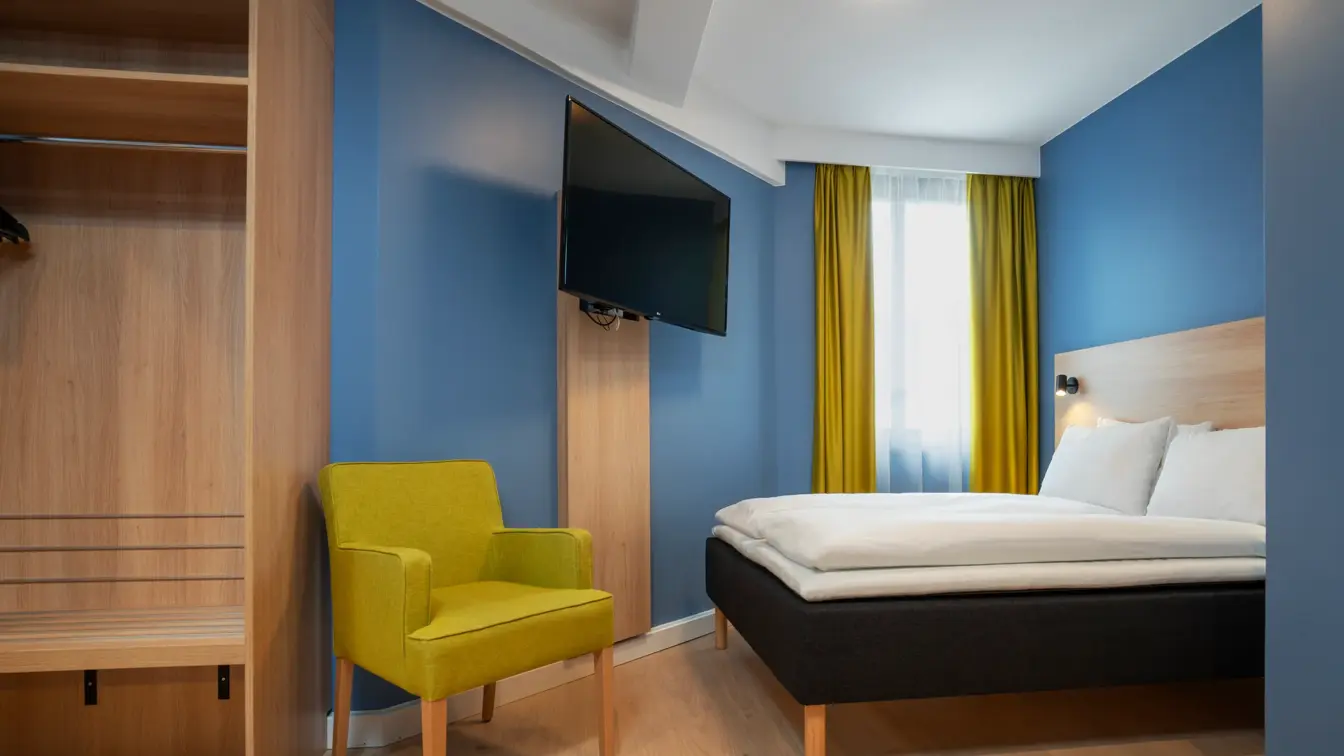 Garderobe, gul stol, blå vegger, TV og queen seng på standard rom queen på Thon Hotel Astoria.