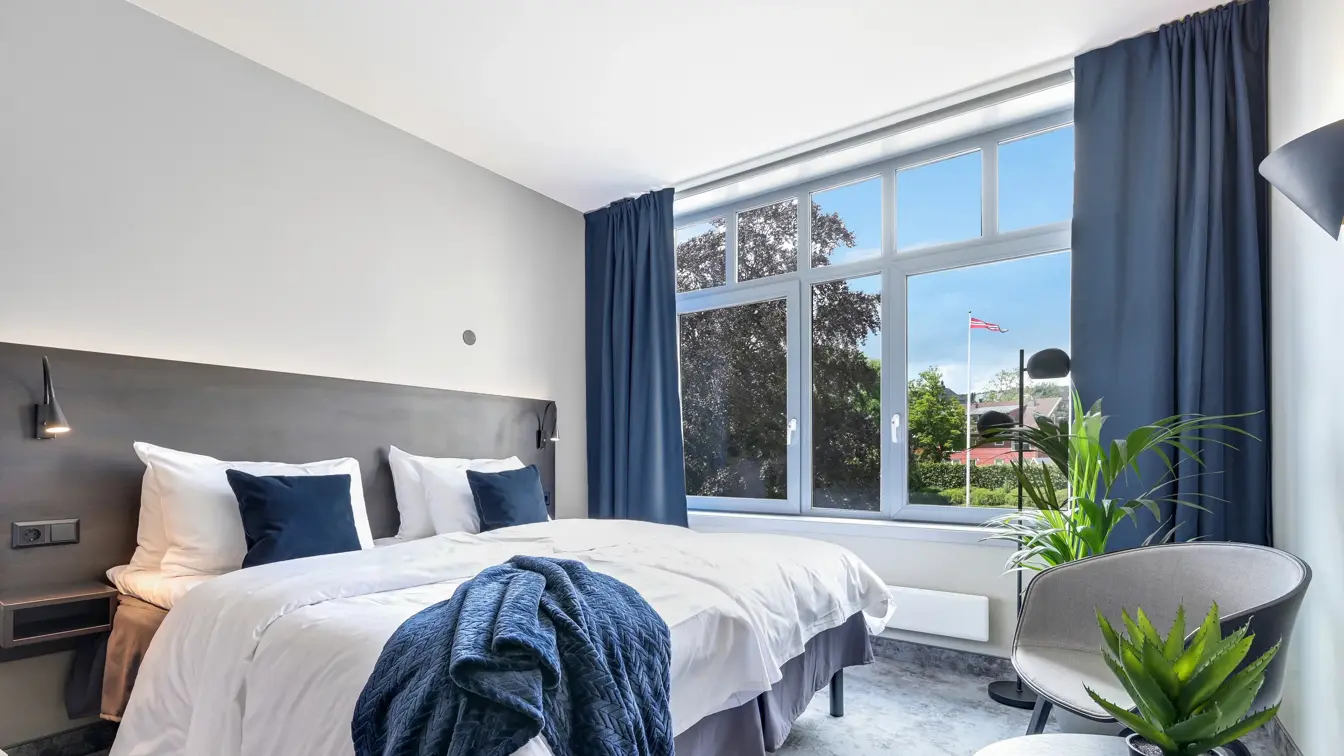 Dobbeltseng, hvite vegger, vindu, blå gardiner, lenestol, salongbord, planter