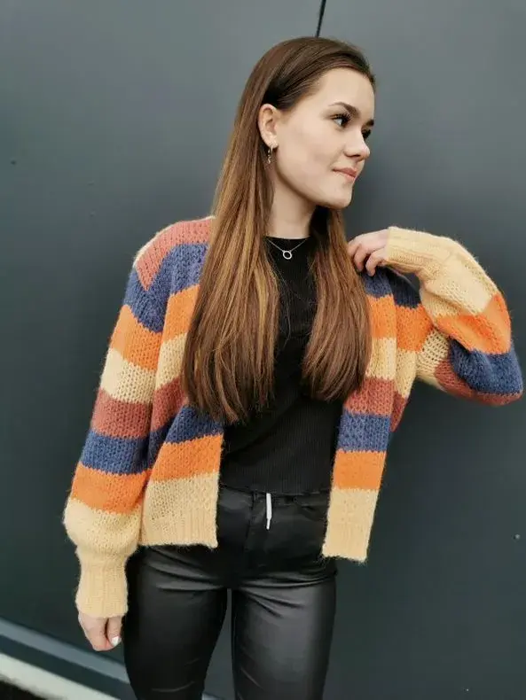 Jente står foran svart vegg. Har mørkt langt hår, svart topp, svart skinnbukse og en fargerik stripete cardigan over. Ser bort fra fotografen. 