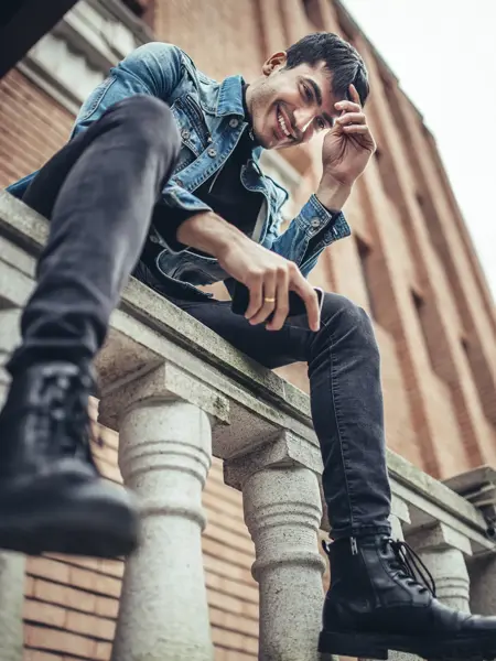 Mann med denimjakke, grå bukse og sorte boots sitter på gjerde sett nedenfra 