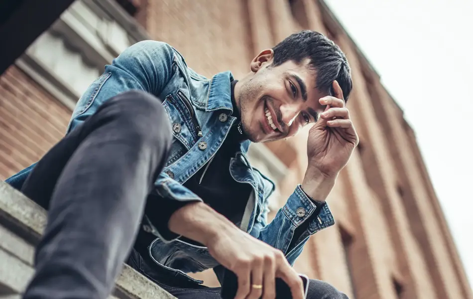 Mann med denimjakke, grå bukse og sorte boots sitter på gjerde sett nedenfra 