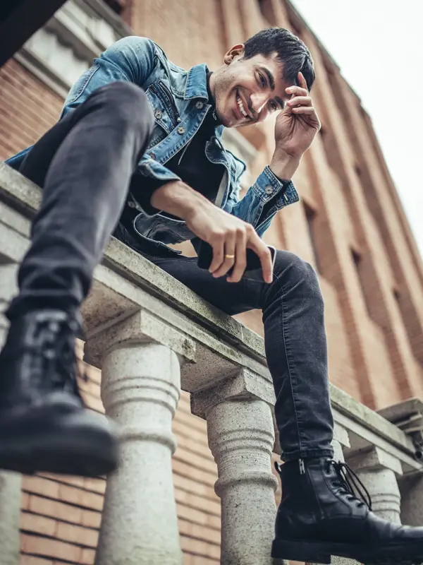 Mann med denimjakke, grå bukse og sorte boots sitter på gjerde sett nedenfra 