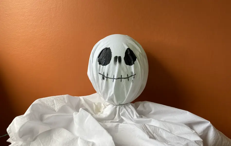 Oppblåst ballong inni et laken med tegnet ansikt som ligger på et bord med oransje bakgrunn