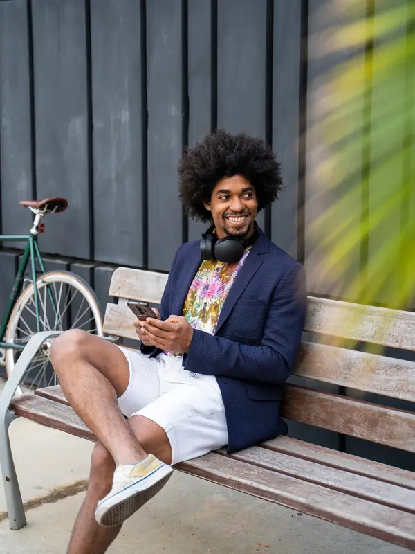 Mann med blå blazerjakke, mønstret t-skjorte og hvit shorts med headset rundt halsen sitter på en benk utendørs og smiler med en palme i forgrunnen 