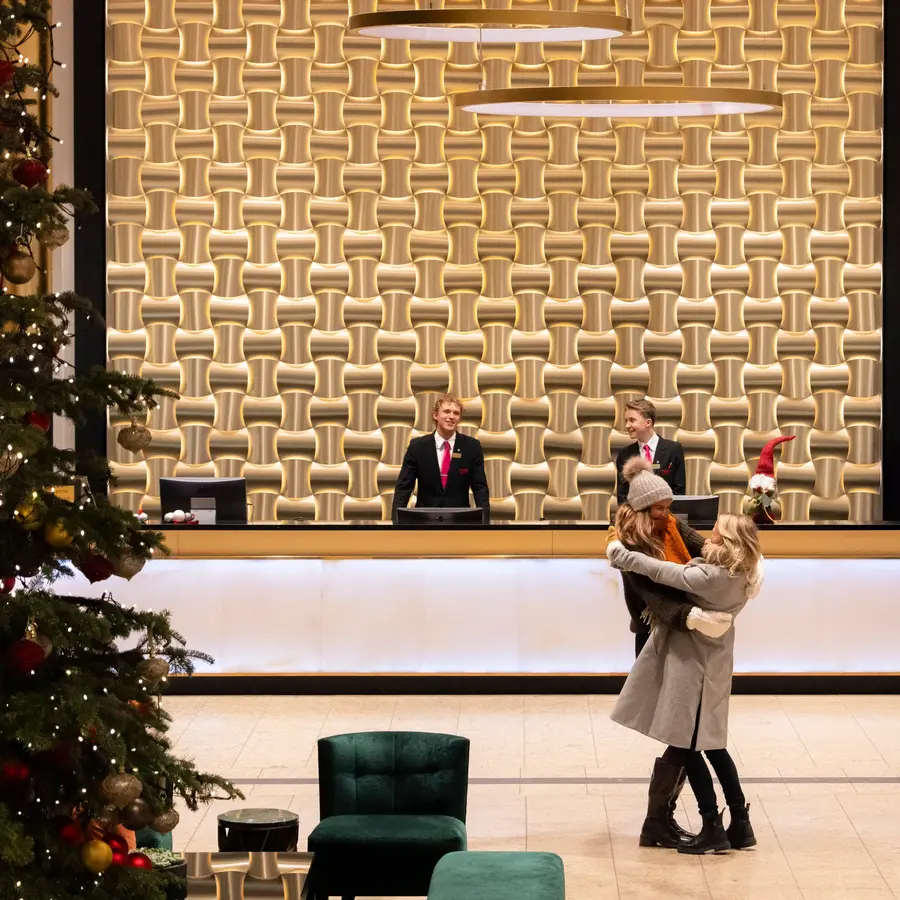 Stort og lysende juletre i lobby, med to personer med ytterklær som klemmer hverandre foran resepsjonsdisk.