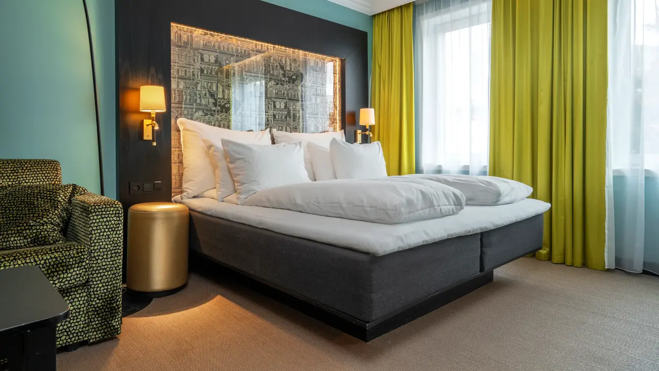 Businessrom med dobbeltseng, turkise vegger, gule gardiner, tente sengelamper og mønstrete lenestol.