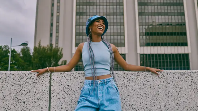 Dame med lys blå topp, lys jeans og blå bøttehatt lener seg mot murvegg og smiler 