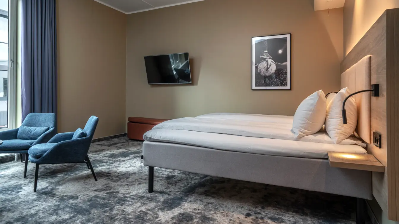 Handicapvennlig rom med dobbeltseng, to blå lenestoler, vegghengt TV og gulvteppe.