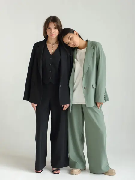 To kvinnelige modeller står inntil hverandre i et hvitt fotostudio. Modellen til venstre er iført svart dress. Modellen til høyre har lysegrønn dress med en hvit t-skjorte under. Modellen til høyre lener hodet sitt på skulderen til modellen til venst