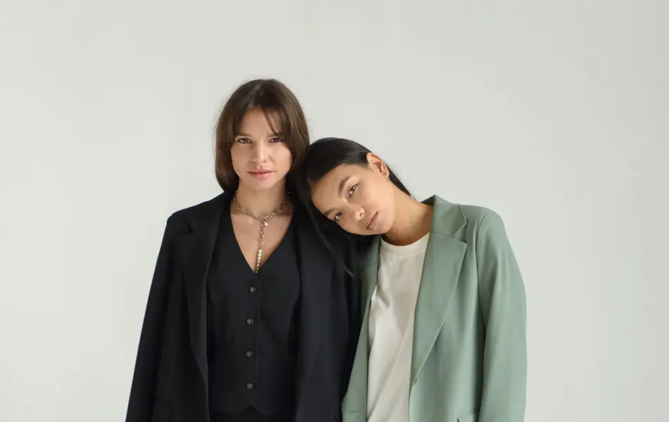 To kvinnelige modeller står inntil hverandre i et hvitt fotostudio. Modellen til venstre er iført svart dress. Modellen til høyre har lysegrønn dress med en hvit t-skjorte under. Modellen til høyre lener hodet sitt på skulderen til modellen til venst