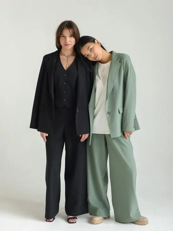 To kvinnelige modeller står inntil hverandre i et hvitt fotostudio. Modellen til venstre er iført svart dress. Modellen til høyre har lysegrønn dress med en hvit t-skjorte under. Modellen til høyre lener hodet sitt på skulderen til modellen til venst