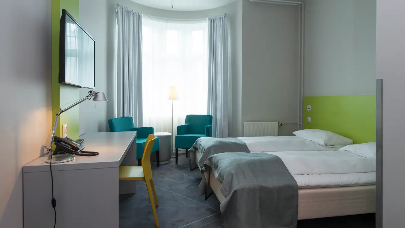 Standard Room Twin på Thon Hotel Trondheim. Lyst rom med store vinduer. Grått teppe, hvit vegg med limegrønne detaljer, to turkise lenestoler, to enkeltsenger, skrivebord med tilhørende gul stol. TV på veggen og stort speil.