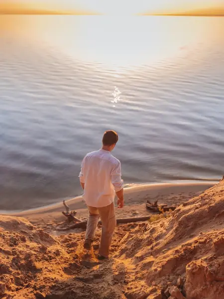 Mann i hvit linskjorte går ned til vannet på en strand i solnedgang 