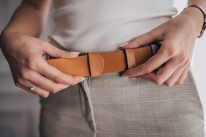 Nærbilde av hender som strammer et belte i brunt semsket skinn