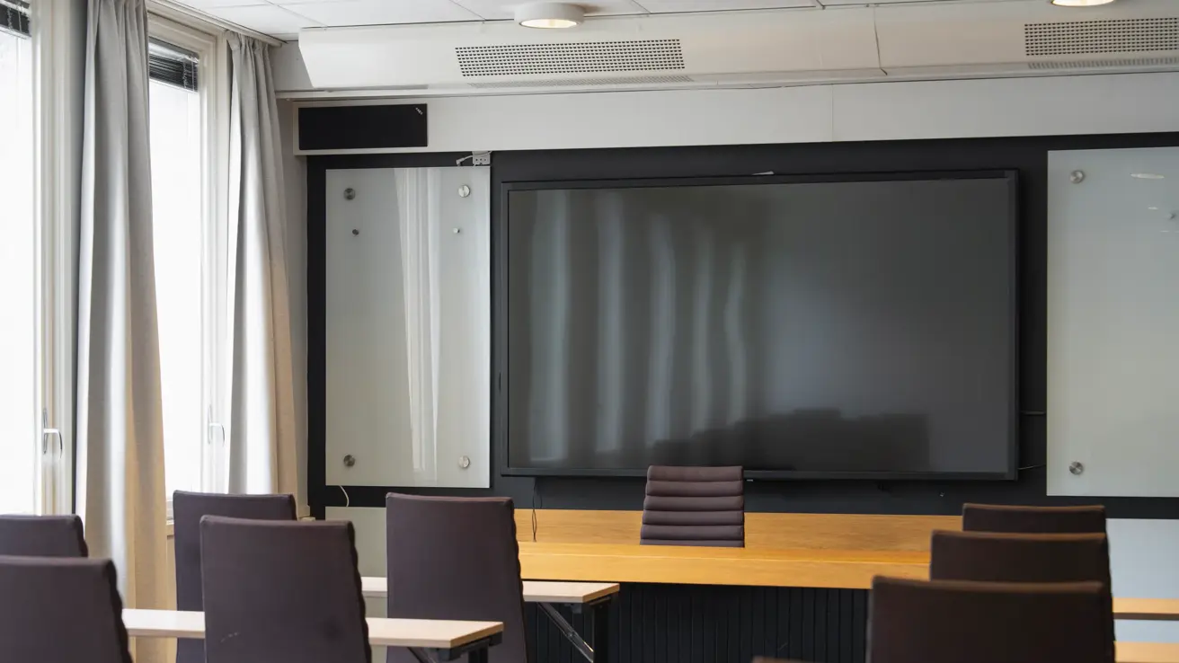 Nærbilde av noen bord og stoler i møterom med stor TV skjerm foran