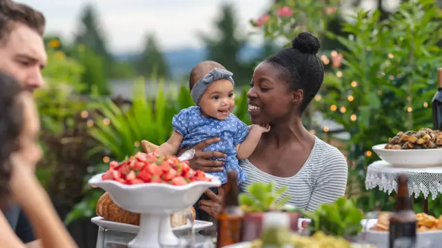 A multiethnic group of young friends enjoy good food and conversation together on a terrace outside on a summer evening.  The focus is on an African American mother who is holding her infant daughter in her lap.
