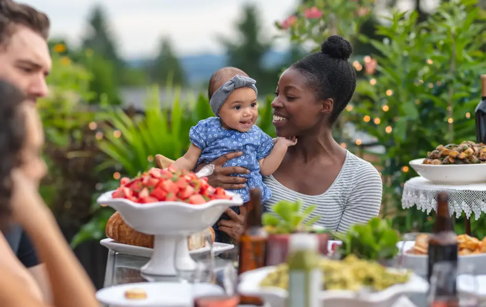 A multiethnic group of young friends enjoy good food and conversation together on a terrace outside on a summer evening.  The focus is on an African American mother who is holding her infant daughter in her lap.