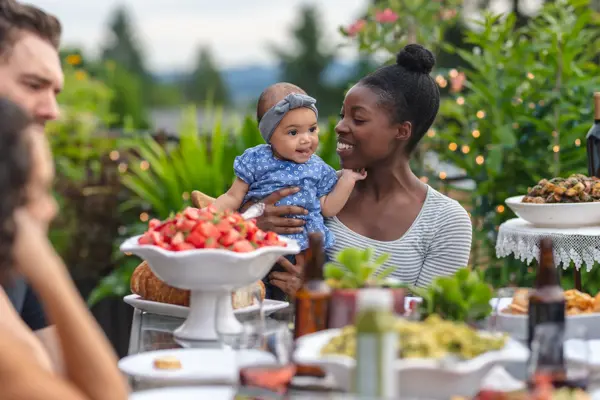 A multiethnic group of young friends enjoy good food and conversation together on a terrace outside on a summer evening.  The focus is on an African American mother who is holding her infant daughter in her lap.