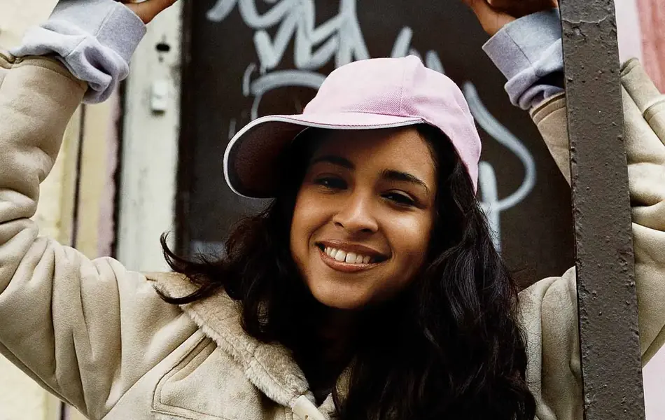 Dame med stor jakke i lyst skinnaktig stoff og med lys caps, står bak et rekkverk i metall og ser ned mot kamera.