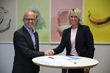 Dame og mann utveksler håndtrykk ved et bord med papirer, vegg med fargerike bilder av mat i bakgrunnen.