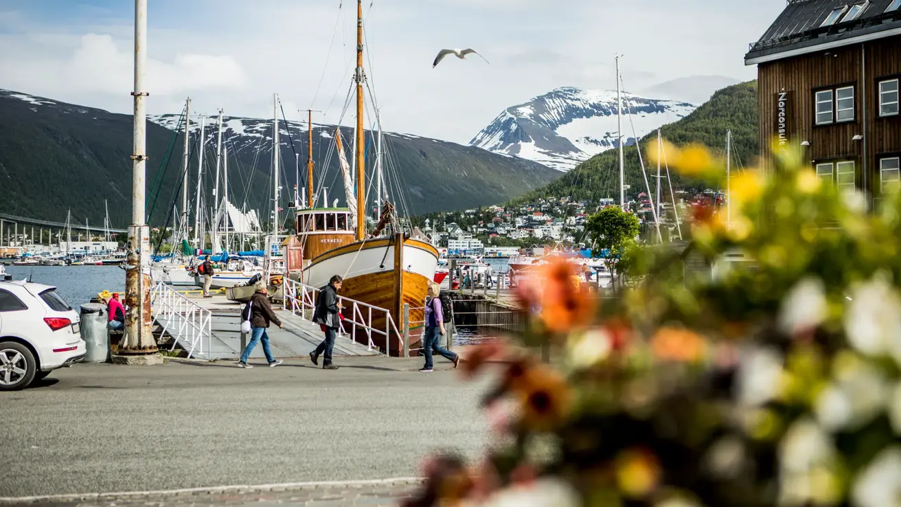 En fiskebåt ligger fortøyd ved en av bryggene i Tromsø. I bakgrunnen ses Ishavskatedralen, grønne fjell og noen fjell med snø.