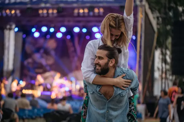 Mann i denimskjorte med en kvinne på ryggen. Kvinnen har armen i været. I bakgrunnen ser man en scene.