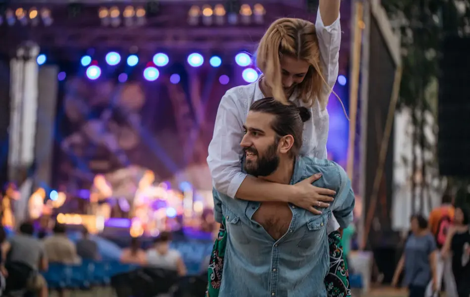 Mann i denimskjorte med en kvinne på ryggen. Kvinnen har armen i været. I bakgrunnen ser man en scene.