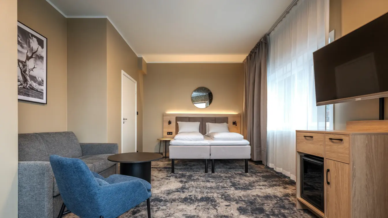 Junior suite med dobbeltseng, grå sofa, blå lenestol, lite sort bord, gulvteppe og TV med TV benk under og minibar.