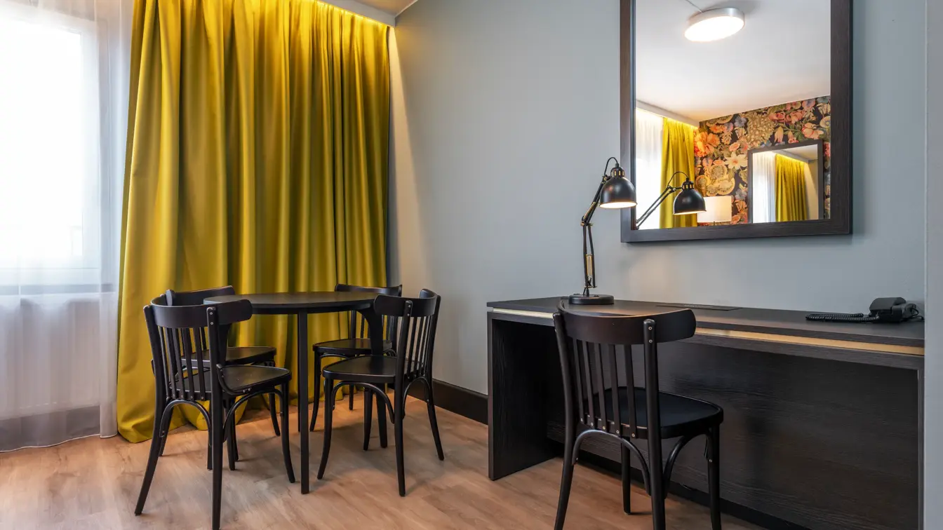 Business rom med rundt spisebord med stoler rundt, skrivepult, speil på veggen og gul gardin foran vindu.