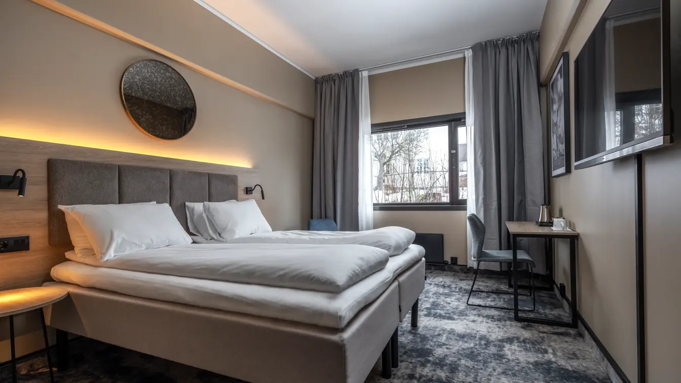 Dobbeltrom med dobbeltseng, hvitt sengetøy, vegghengt TV, gulvteppe, lange grå gardiner og stort vindu i enden av rommet.