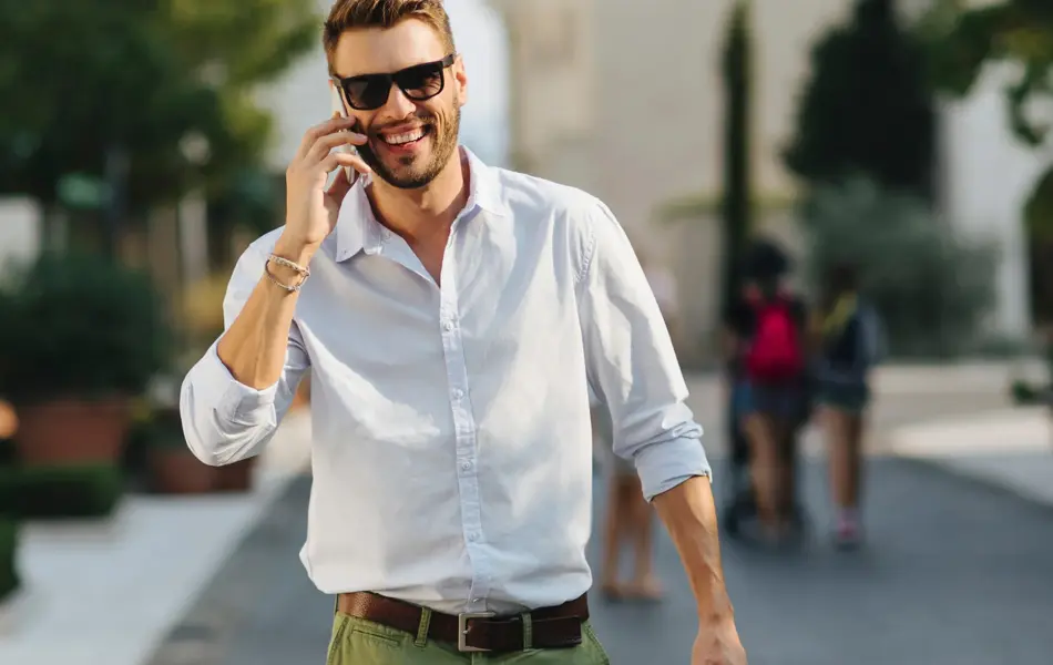Mann med skjorte og shorts går og smiler mot kamera mens han snakker i mobilen 