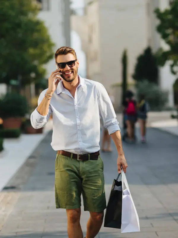 Mann med skjorte og shorts går og smiler mot kamera mens han snakker i mobilen 