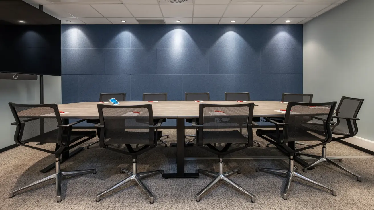 Møterom med styrebords oppsett til 10 personer, spotter i taket, en blå vegg, TV skjerm, penn og papir på bordet.