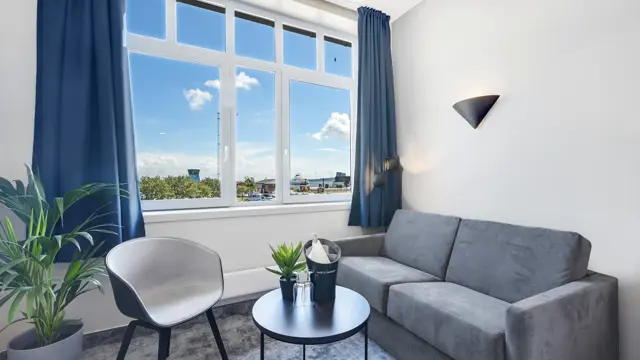 HVite vegger, vindu, blå gardiner, grå sofa, lenestol, salongbord, planter 