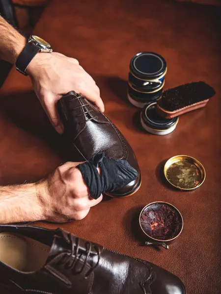 Nærbilde av hender som pusser sko, med diverse skopusserutstyr på bord med dyp rød overflate. Foto til artikkel om gavetips til farsdag.