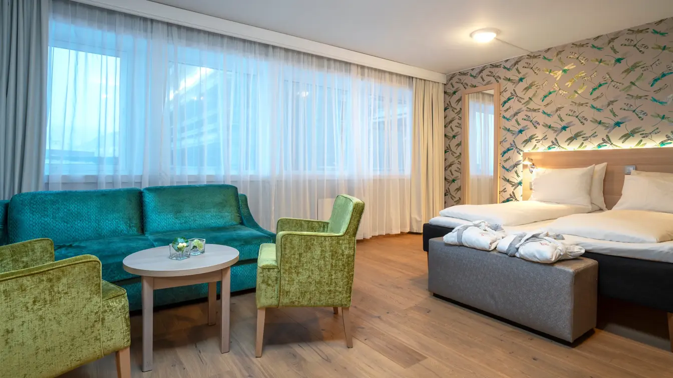 Fargerik tapet, store vinduer, sittegruppe med grønn sofa, gule stoler og bord og dobbeltseng med morgenkåper i business rom på Thon Hotel Polar i Tromsø.