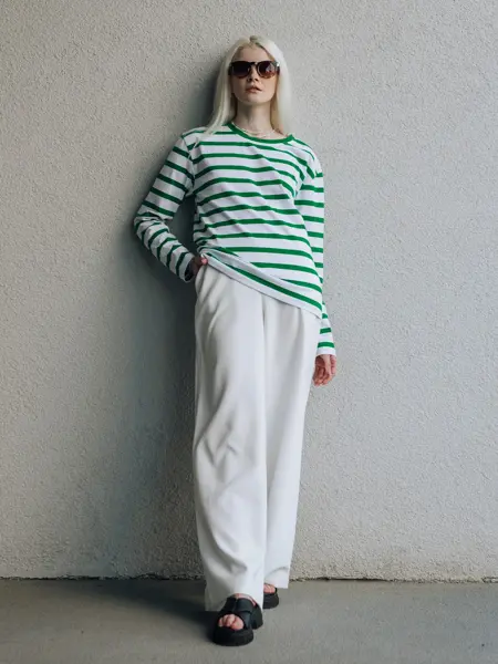 Dame med solbriller, hvit, løs bukse og grønn og hvitstripete genser poserer inntil hvit murvegg. 