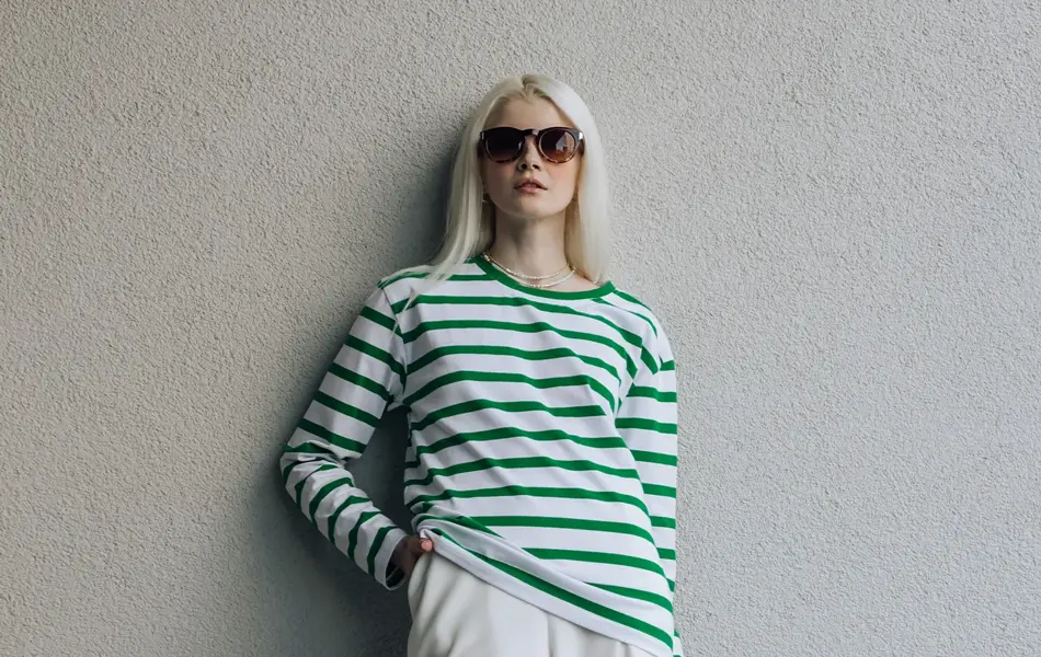 Dame med solbriller, hvit, løs bukse og grønn og hvitstripete genser poserer inntil hvit murvegg. 