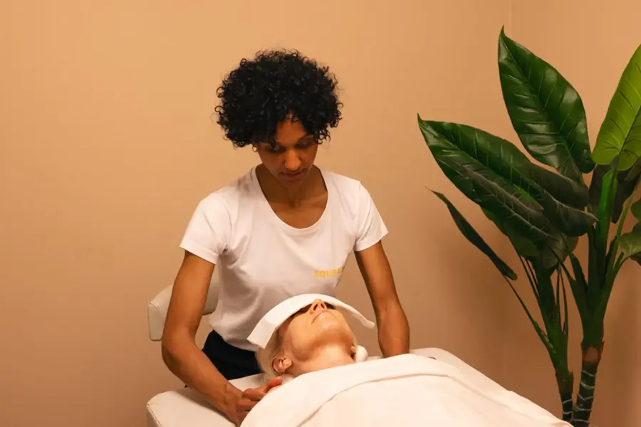dame med krøllete sort hår som gir en annen dame med massasje på massasje seng med grønt plante ved siden av rommet