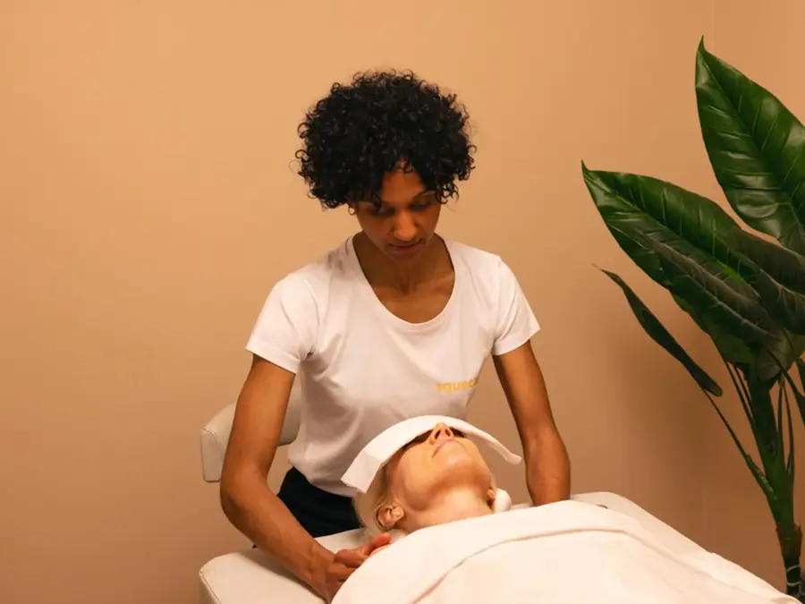 dame med krøllete sort hår som gir en annen dame med massasje på massasje seng med grønt plante ved siden av rommet