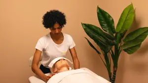 dame med krøllete sort hår som gir en annen dame med massasje på massasje seng med grønt plante ved siden av rommet