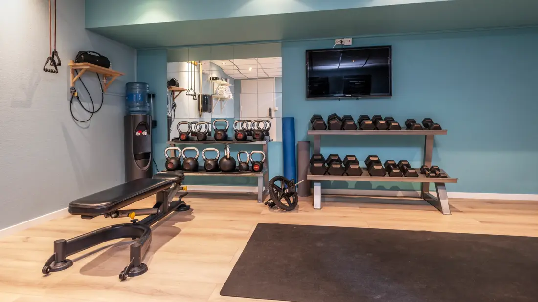 Treningsrom med benk, trenings matte på gulvet, manualer på hyller, TV på veggen.