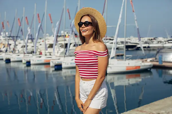 Dame med kort hvit shorts og stripete t-skjorte smiler foran båtmarina